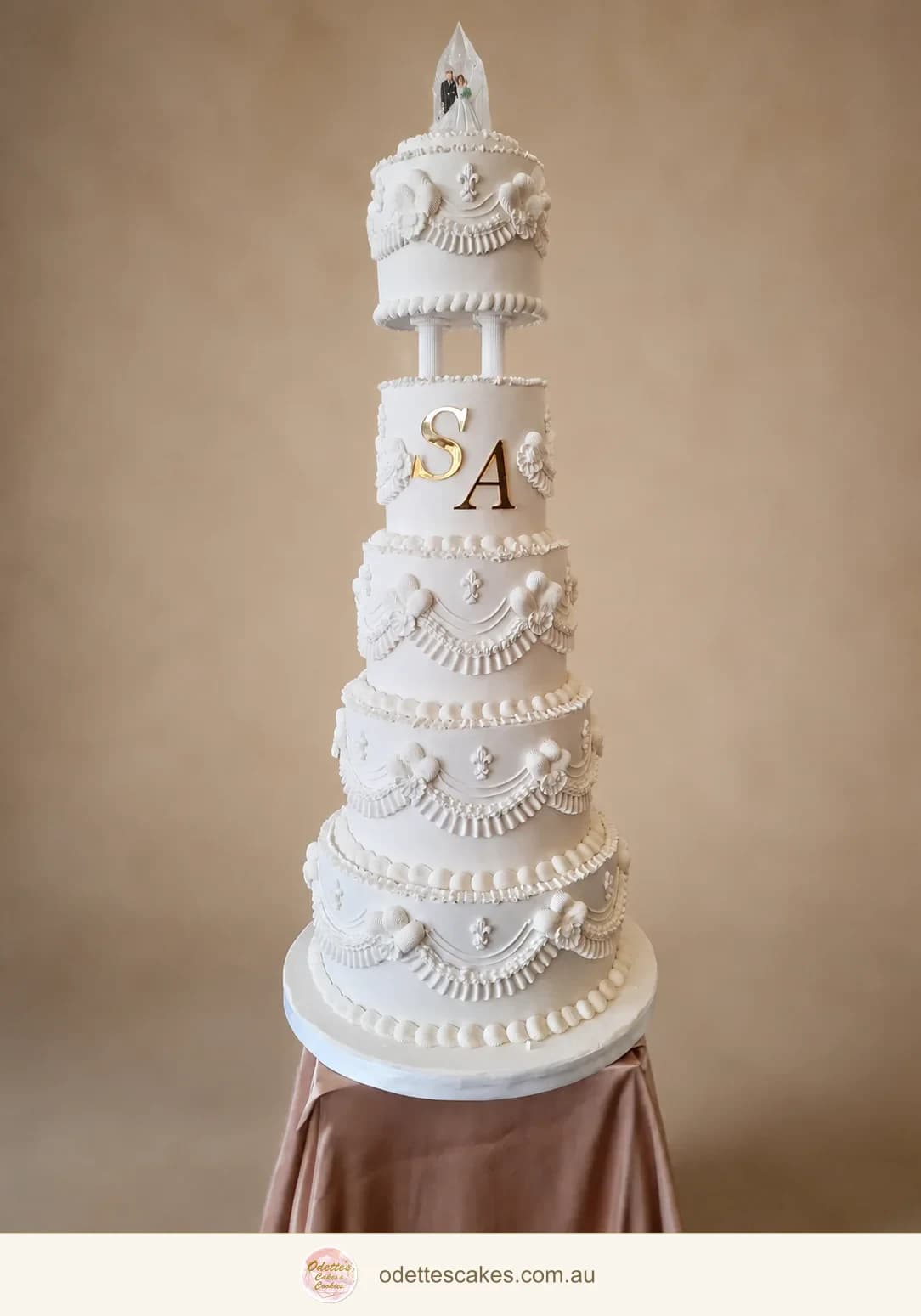 Tall five-tier white wedding cake with ornate piped swags, floral details and gold initials