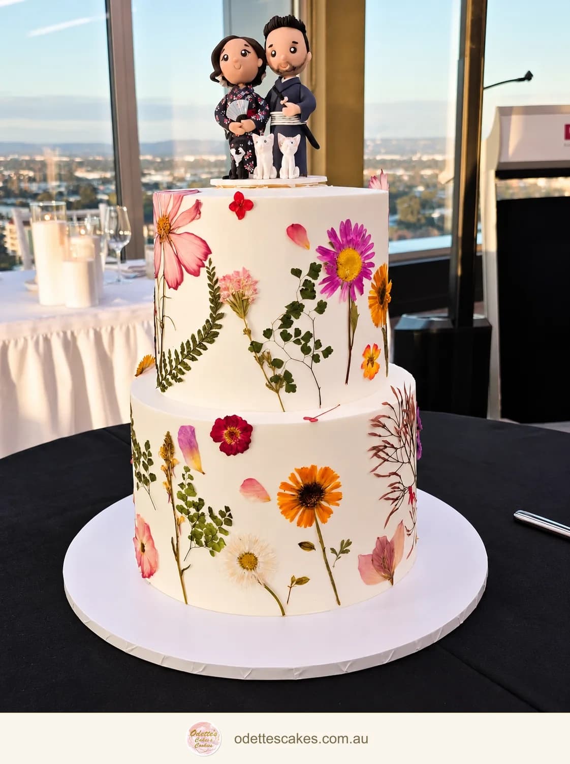 Two-tier white wedding cake with colourful pressed flowers and a personalised couple and cats topper, by Odette in Jindalee, Perth