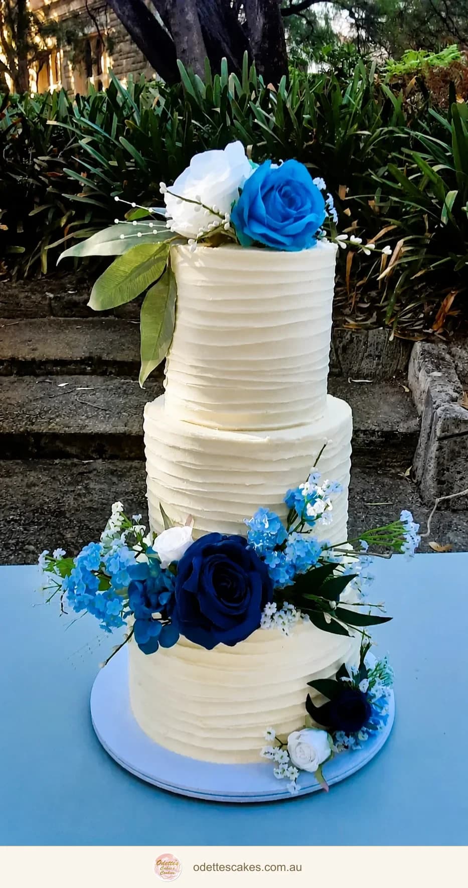 Three-tier ivory wedding cake with royal blue roses and white flowers at an outdoor venue